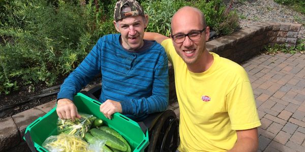 Two friends smiling at camera, one kneeling and the other in a wheelchair holding a box of veggies