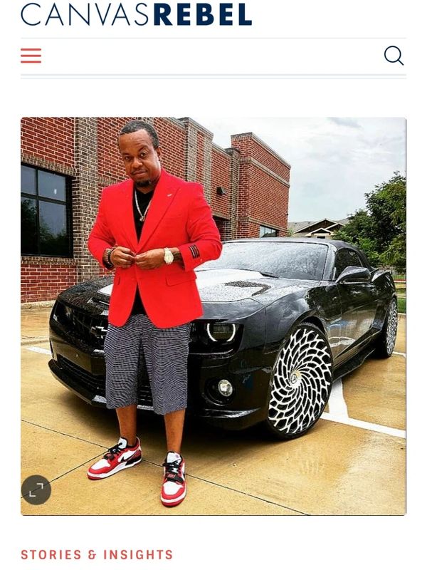 Man in red blazer and sneakers posing by a black sports car with unique rims.