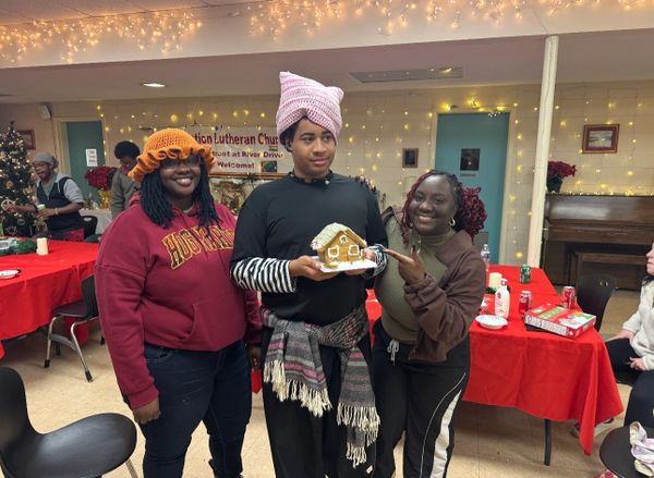 Three friends proudly display a gingerbread house at a festive holiday gathering.
