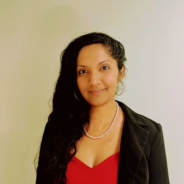 Woman with long dark hair wearing a red top and black blazer, smiling gently.