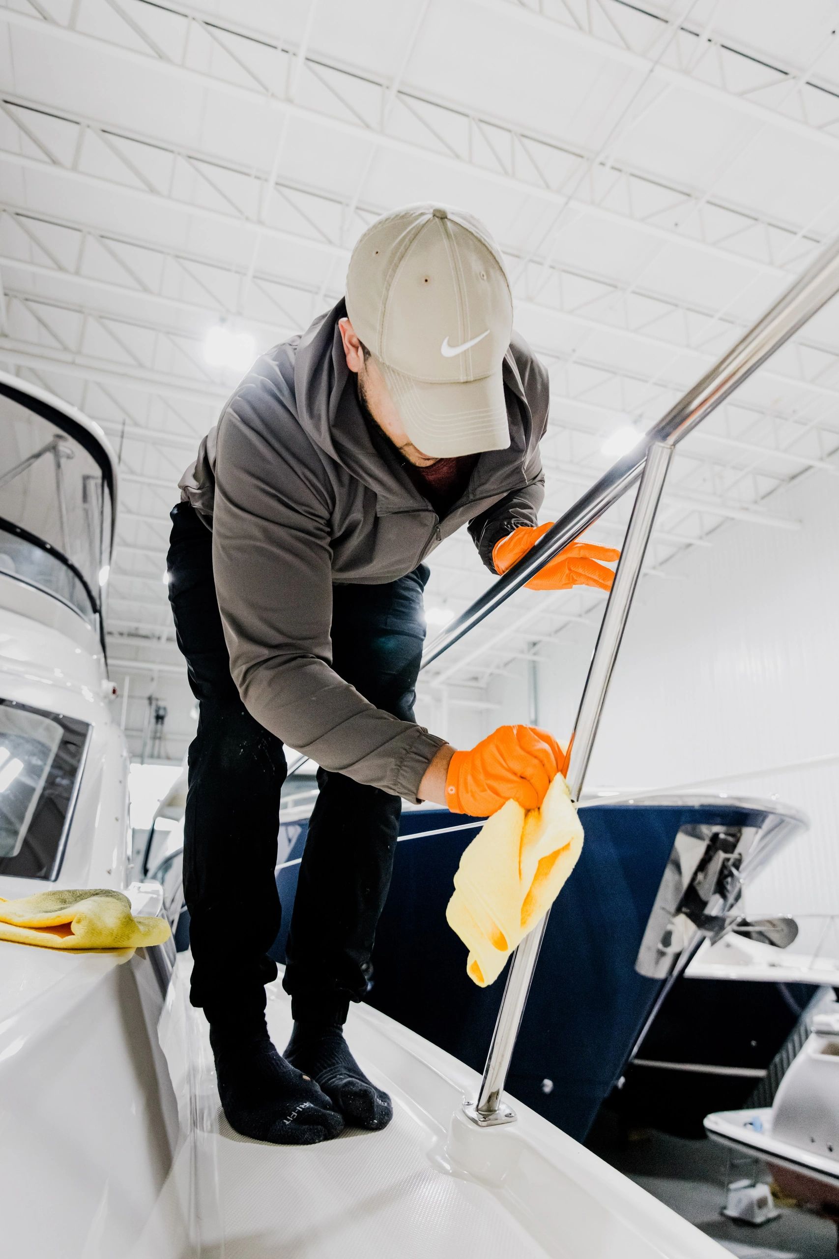 A marine detailer wearing a cap and orange gloves wipes stainless steel railings on a boat inside a 