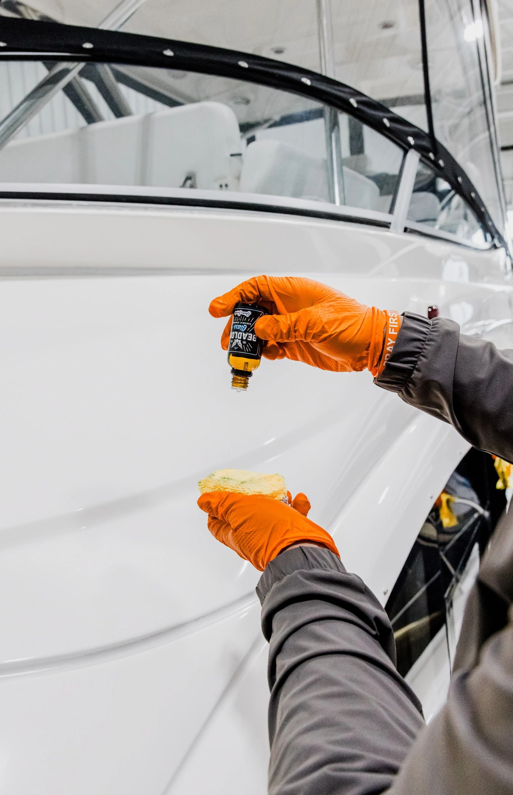 Gloved hands apply a ceramic coating product to the smooth white surface of a boat hull inside an in