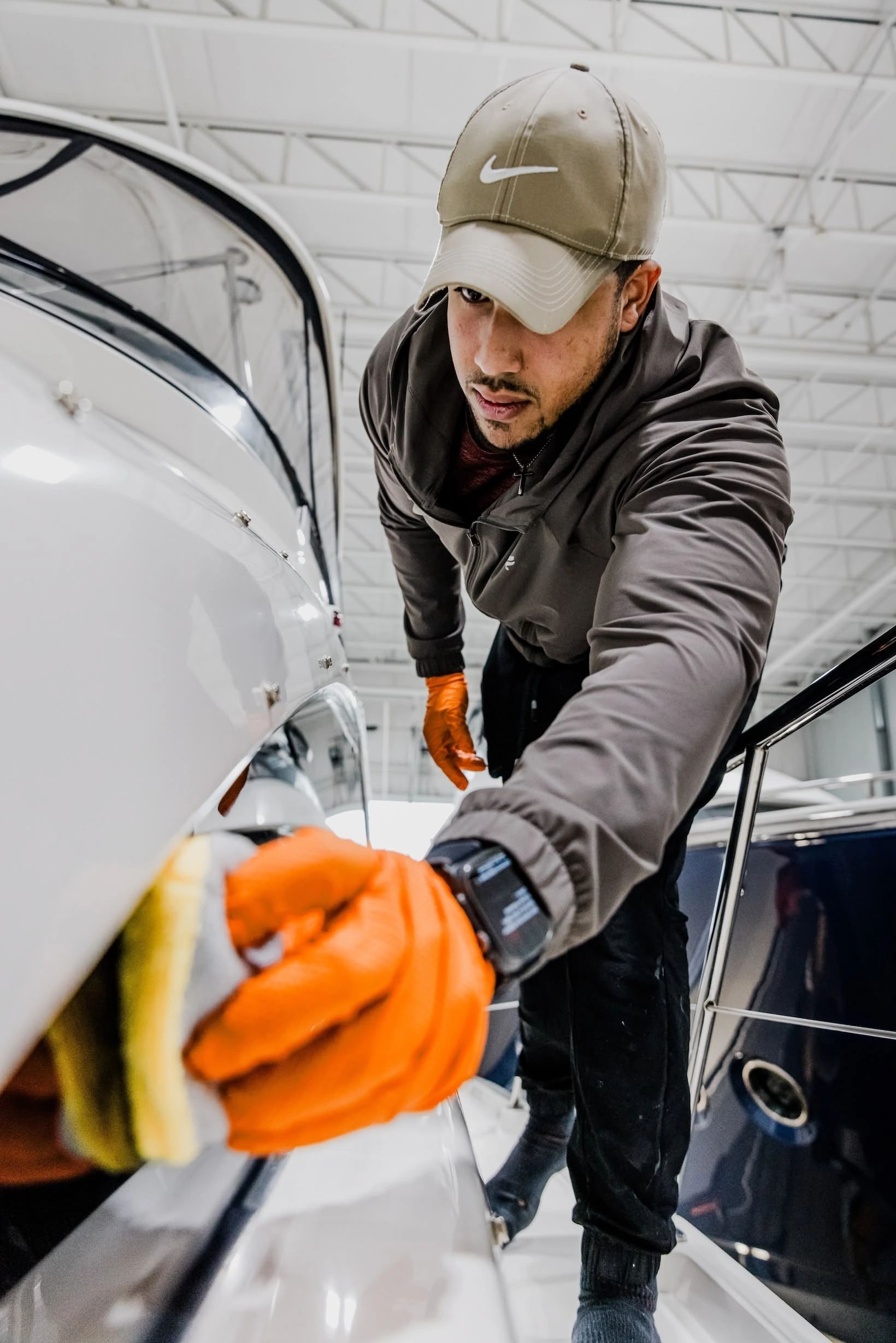 A marine detailer wearing a cap and orange gloves carefully polishes the glossy white surface of a b