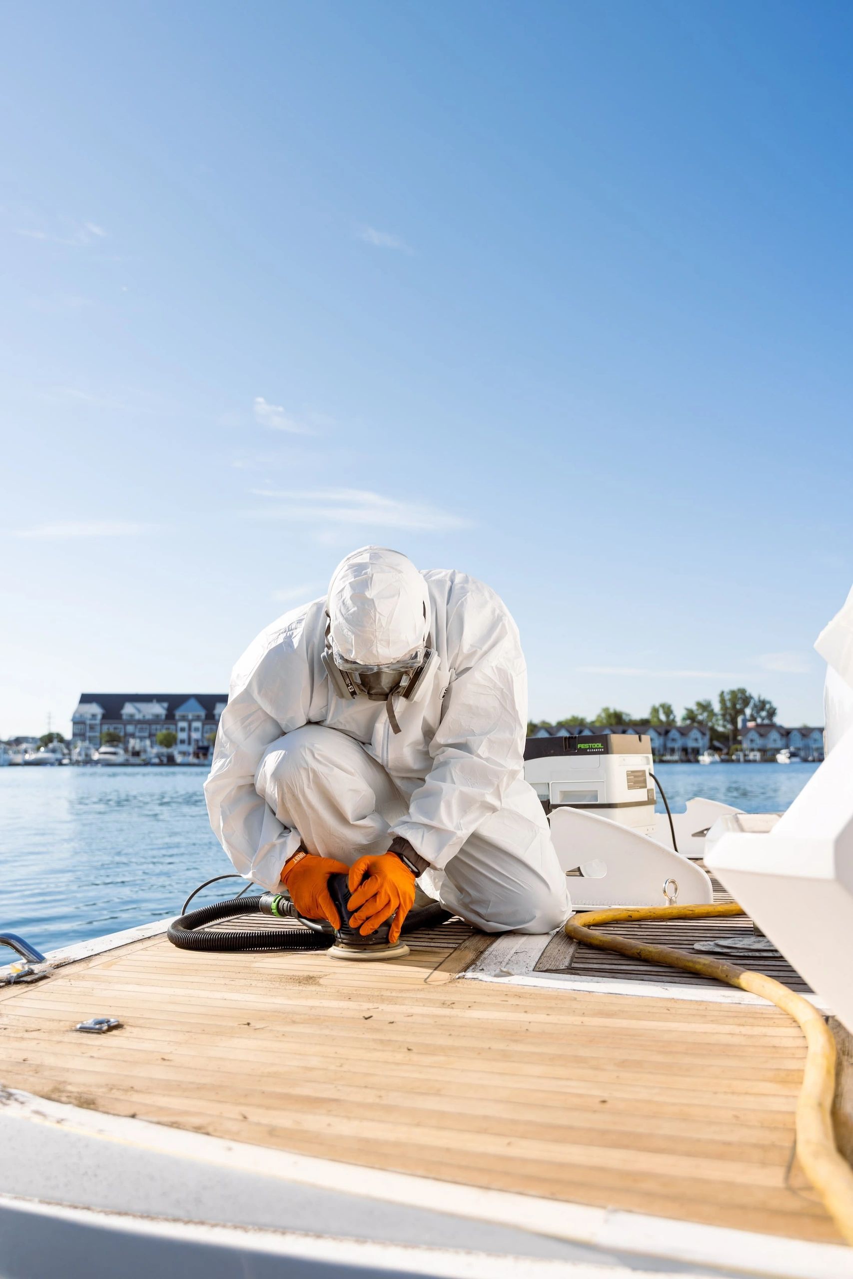 A technician wearing white protective gear and orange gloves kneels on the wooden deck of a yacht, w