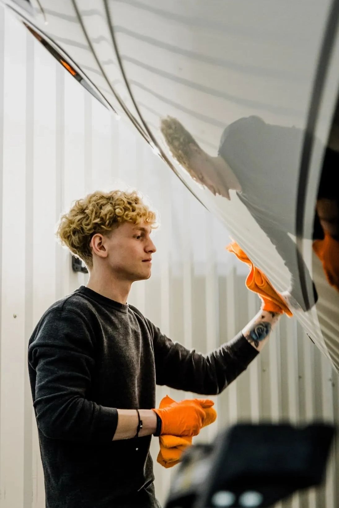 A technician wearing orange gloves works carefully along the side of a boat hull inside an indoor se