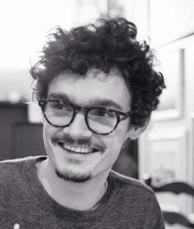 A young man with curly hair and glasses smiling warmly.