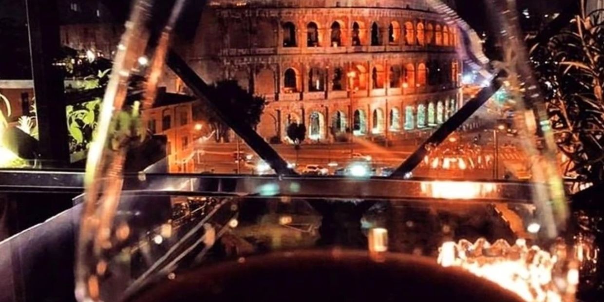 Night view of the Colosseum in Rome from a terrace and trough a wine glass with some red wine in it.