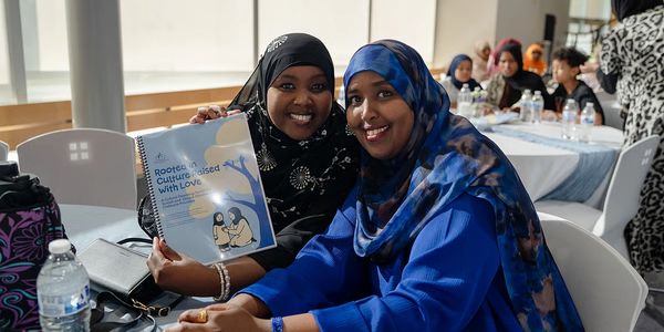Two women smiling and holding a book titled 'Rooted &amp; Culture Raised with Love'.