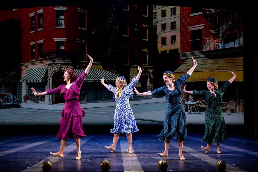 Four women in vintage dresses joyfully dancing on stage with a city backdrop.