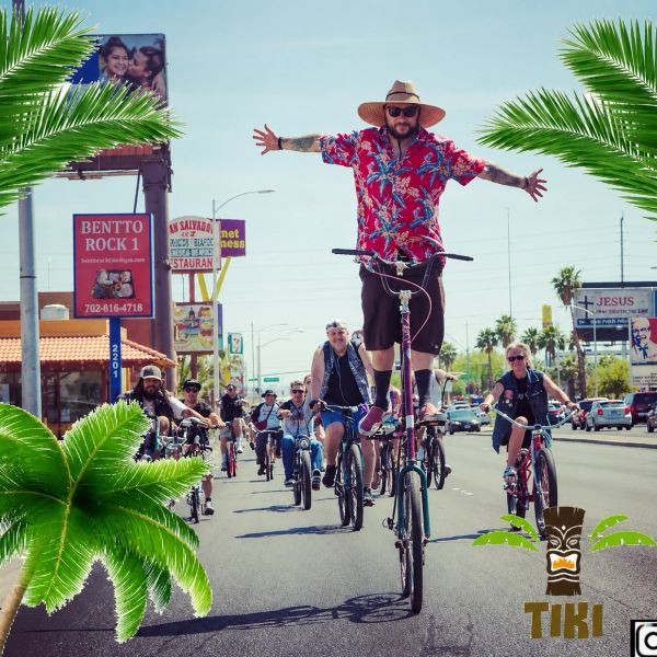 Man in a red Hawaiian shirt rides a tall bike with arms outstretched in a group bike ride.