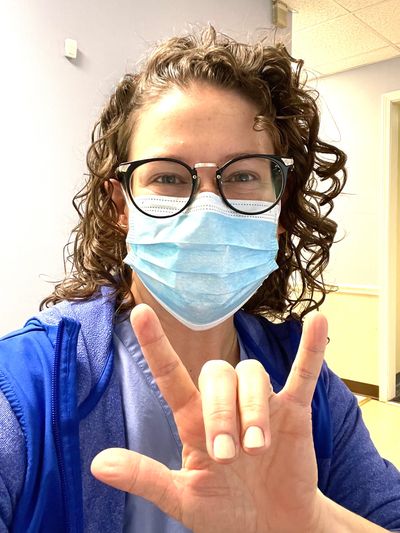 Dr. McHugh is wearing a mask, eyes smiling, and hand holding up an "I love you" sign.