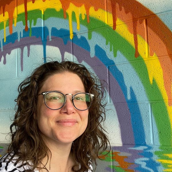 Katie McHugh, MD stands in front of a concrete wall with a rainbow painted on it. 