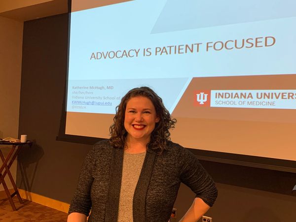 Dr. McHugh stands smiling in front of a screen with a slide reading "Advocacy is patient focused".