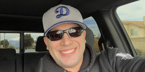 Man wearing sunglasses and a cap smiles inside a car.