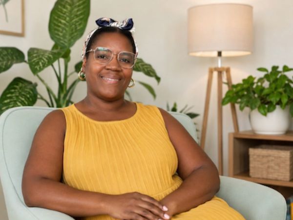 A smiling woman in a yellow dress sitting comfortably in a chair indoors.