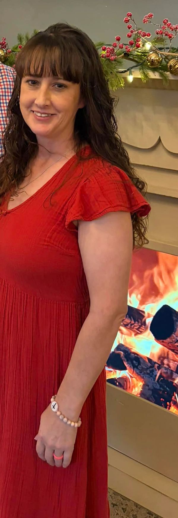 Woman in red dress smiling by a cozy fireplace with holiday decorations.