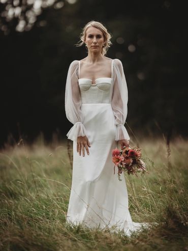 Bride in a white dress with sheer sleeves holding a bouquet in a grassy field.