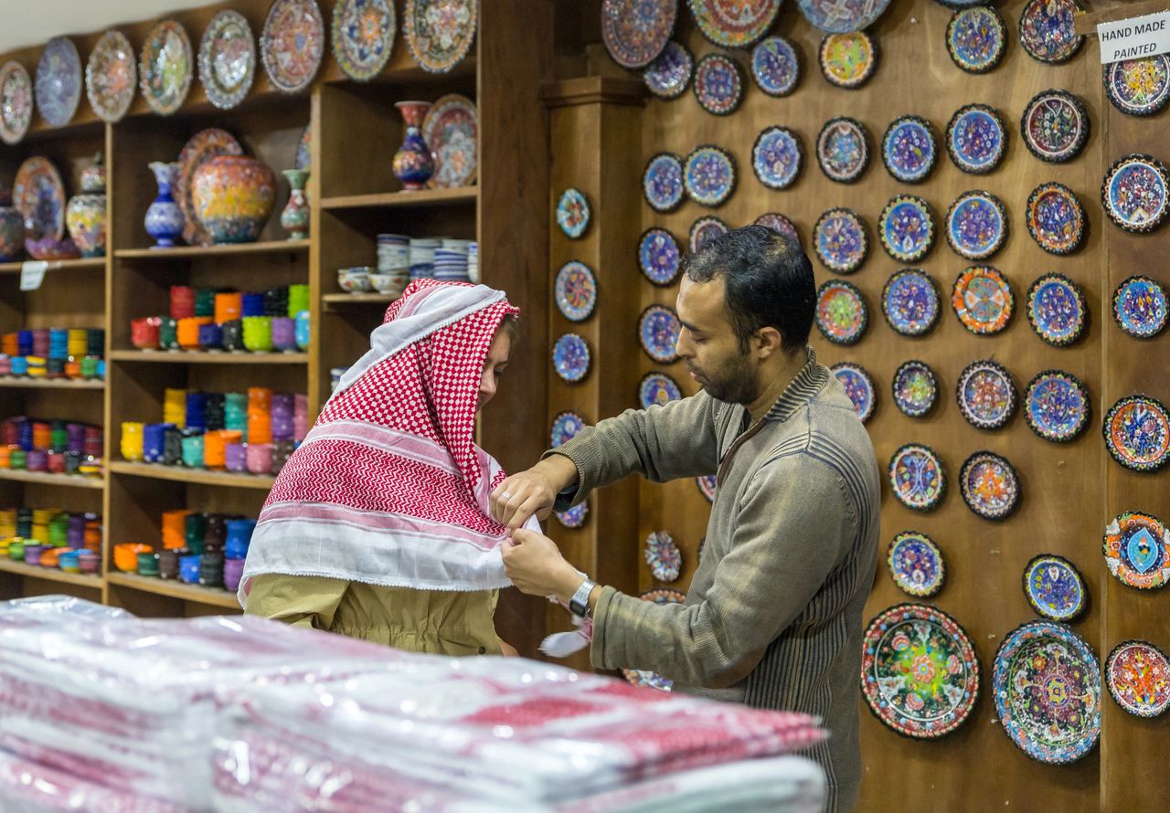 A Middle Eastern artisan assists a customer in selecting a traditional red shemagh scarf from a handmade pottery and textile shop.