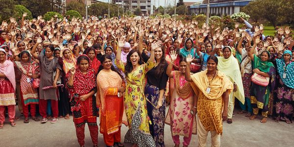 Large group of women in colorful traditional attire waving and smiling outdoors.