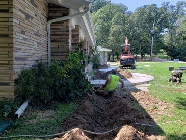 Worker digging a trench beside a house with an excavator nearby.