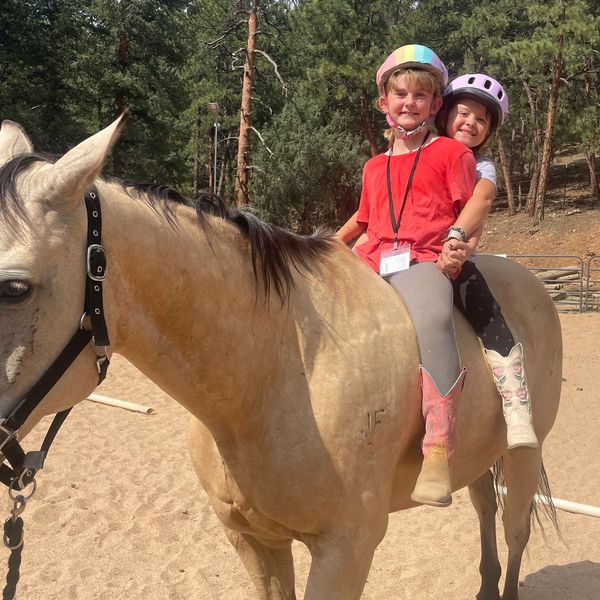 Two children riding a horse with helmets in a sandy arena.