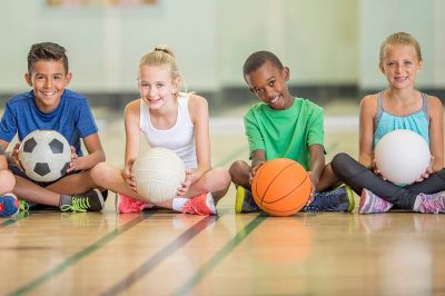 Four kids sitting on gym floor holding different sports balls.