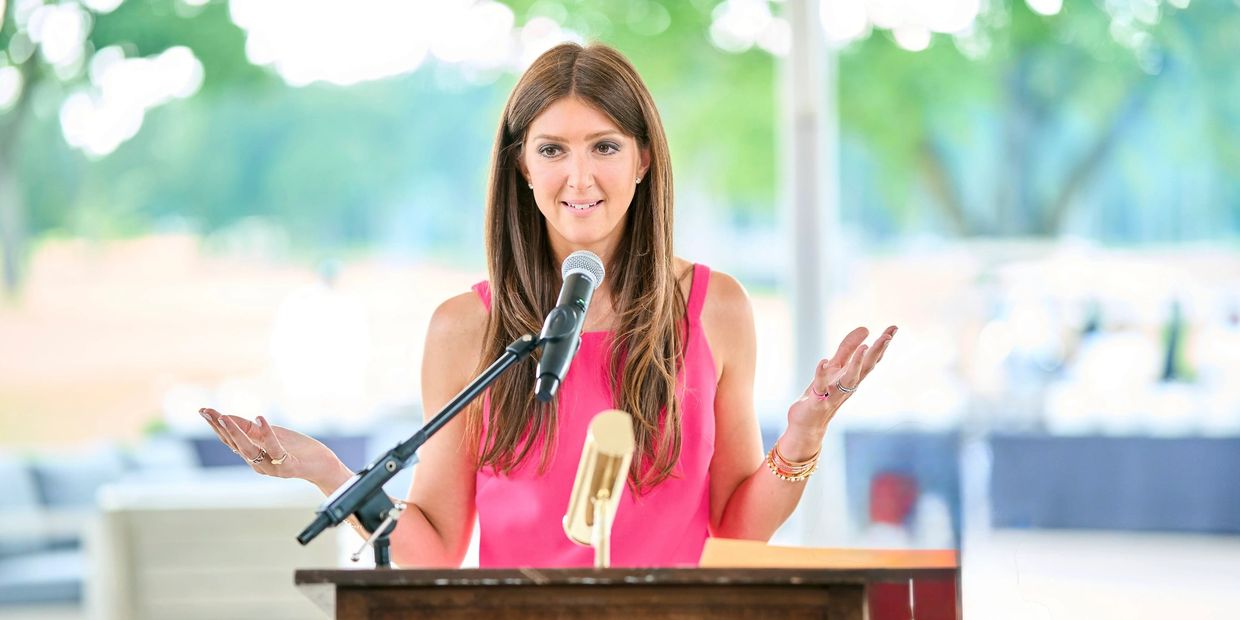 Woman in pink speaking confidently at a podium with microphones outdoors.