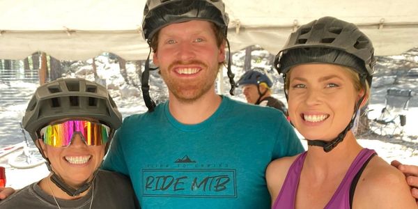 Three smiling cyclists wearing helmets pose together outdoors.