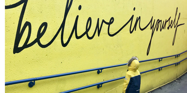 Child in yellow coat looks at 'believe in yourself' mural on bright wall.