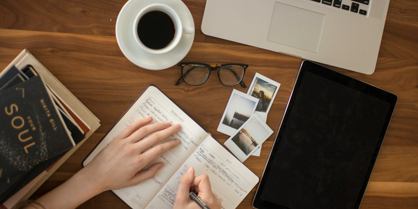 A person writes in a planner surrounded by books, coffee, glasses, and electronic devices on a wooden table.