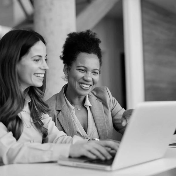 Two women happily providing home buying assistance for women investors, over a laptop at a modern office.