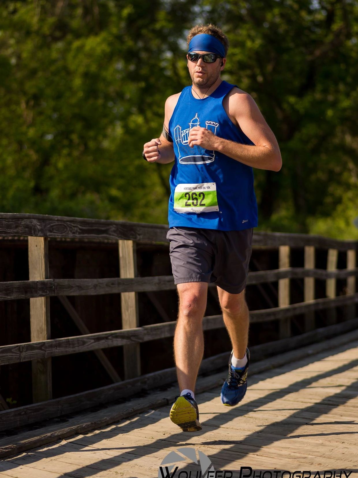 Male runner in blue tank top and sunglasses running on wooden path during race.