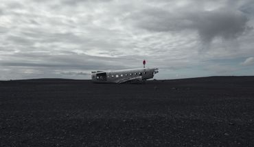 Abandoned airplane wreck on a bleak, dark volcanic landscape under a cloudy sky.