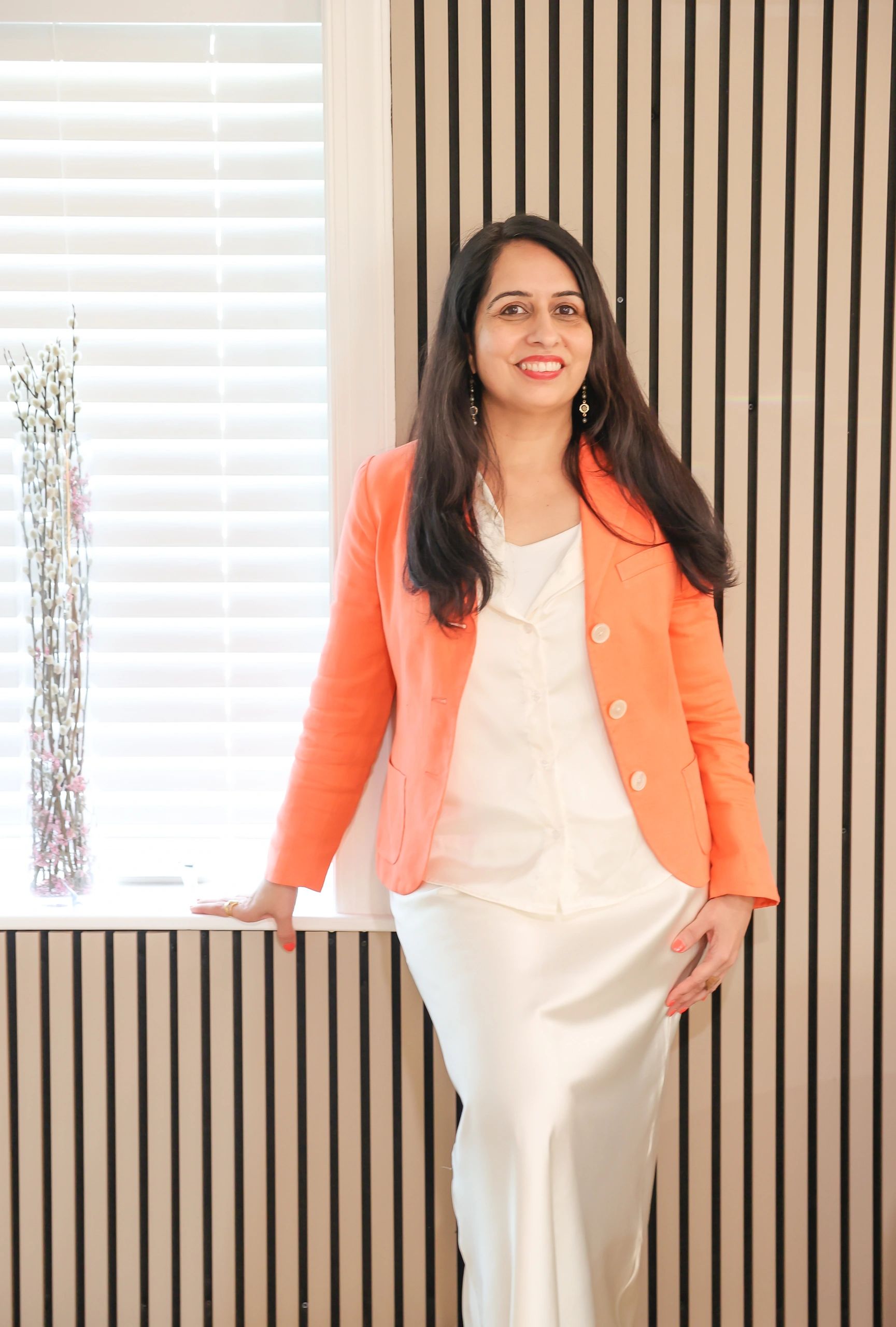 Smiling woman in an orange blazer and white outfit, posing by a window with blinds.