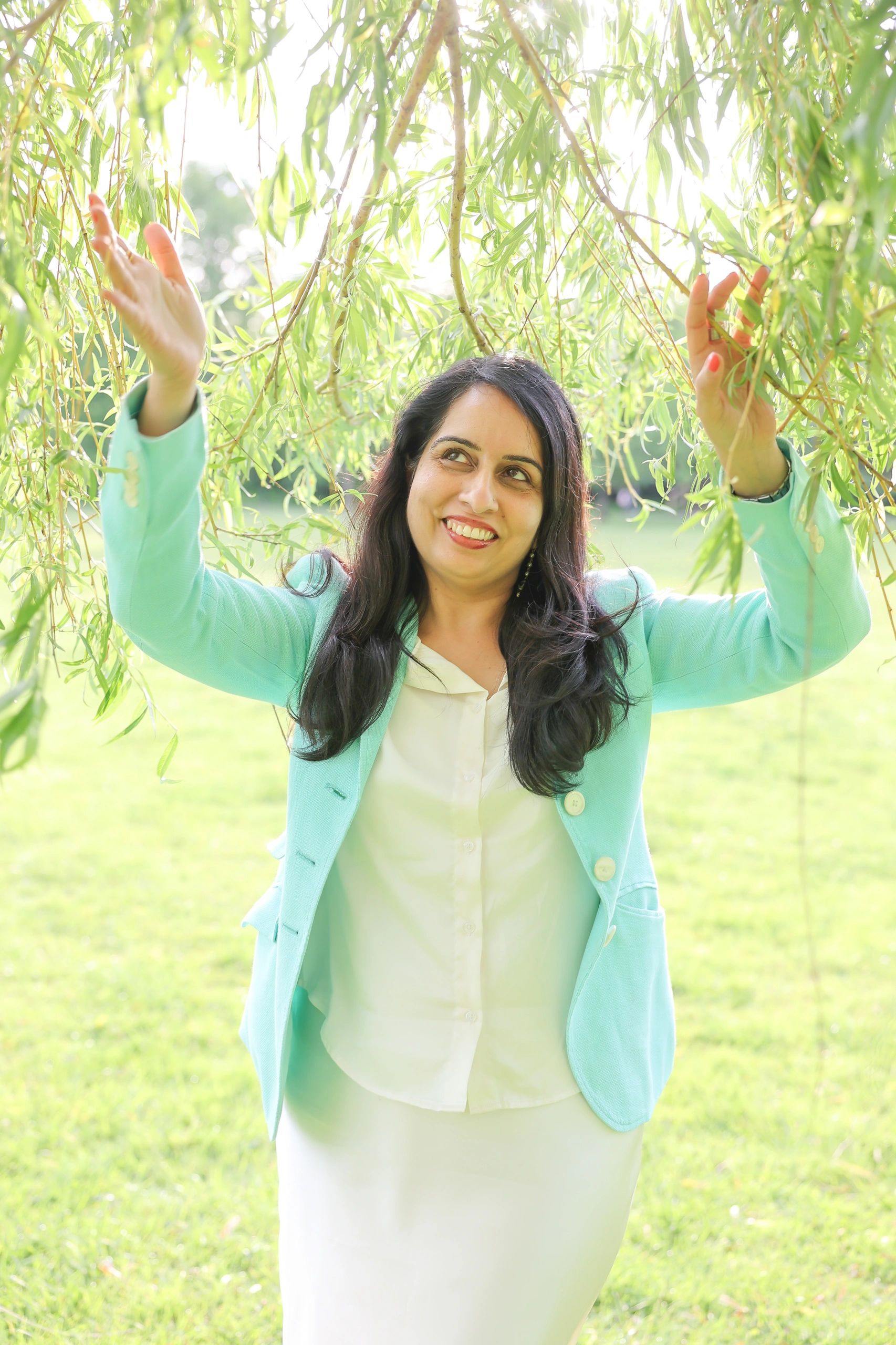 A smiling woman in a light blue blazer enjoys nature under hanging green branches.