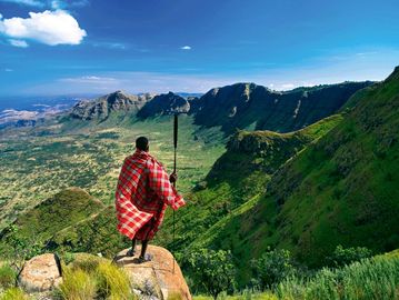 A person in traditional attire overlooking a vast green mountainous landscape.