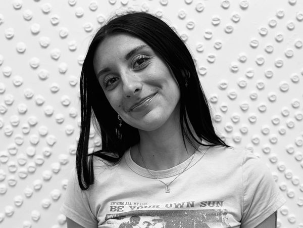 Smiling woman with dark hair wearing a graphic t-shirt against a textured wall.