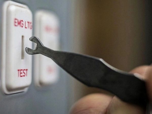 A hand uses a specialized tool to press a test button on a panel labeled EMG LTG.