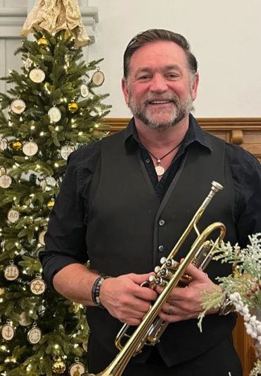 Man smiling with trumpet in front of a decorated Christmas tree.