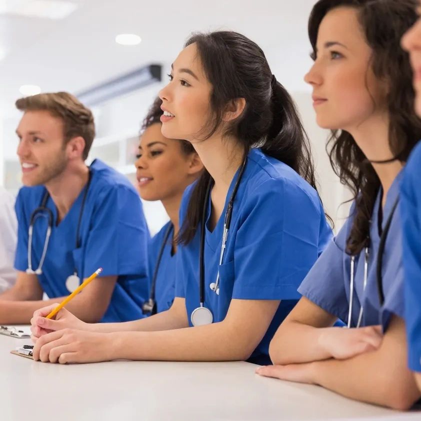 Medical students attentively listen during a lecture, taking notes with stethoscopes around their necks.