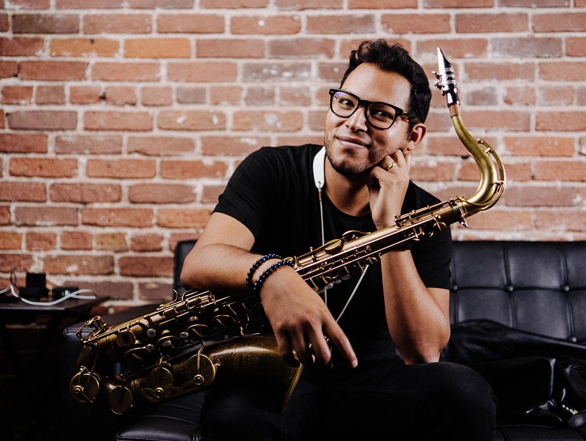 Man with glasses holding a saxophone, sitting against a brick wall.