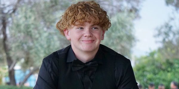 A young man with curly hair dressed in formal black attire smiles outdoors.