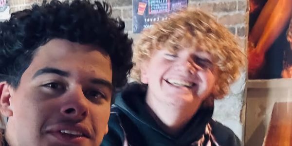 Two young men smiling and posing indoors with brick walls in the background.
