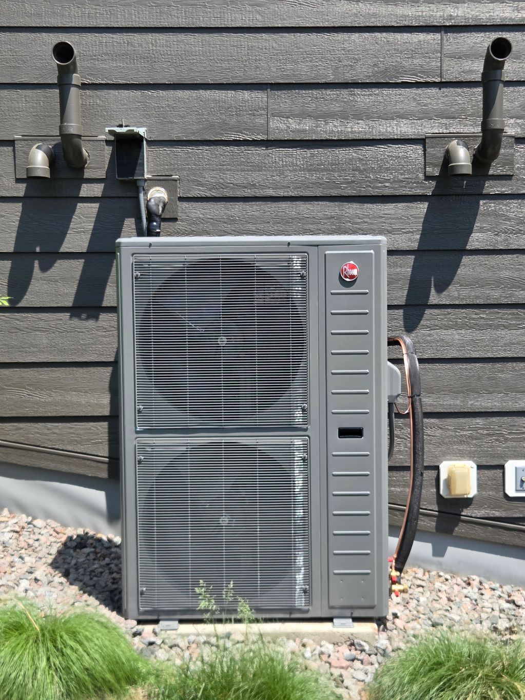 Outdoor HVAC unit installed beside a house with piping and greenery.