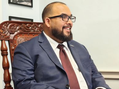 Man in a blue suit and burgundy tie sitting on a carved wooden chair, smiling.
