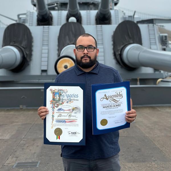 Man holding certificates in front of a large battleship's cannons.
