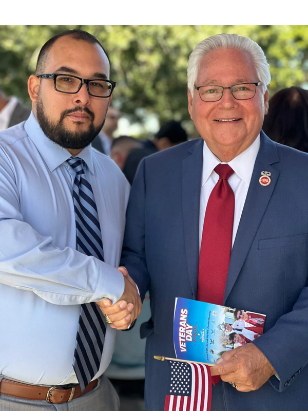 Two men shake hands at a Veterans Day event, one holding a flag and program.