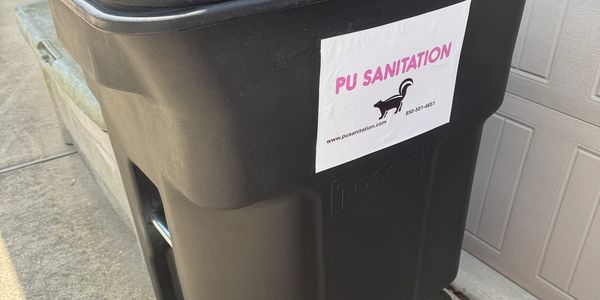 A brown 96-gallon garbage cart with PU Sanitation Logo sticker in front of a garage.