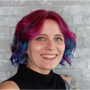 Smiling woman with vibrant pink and blue hair against a gray brick wall.