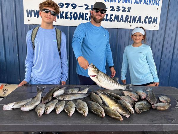 Three people proudly display a large catch of fish on a table at a marina.